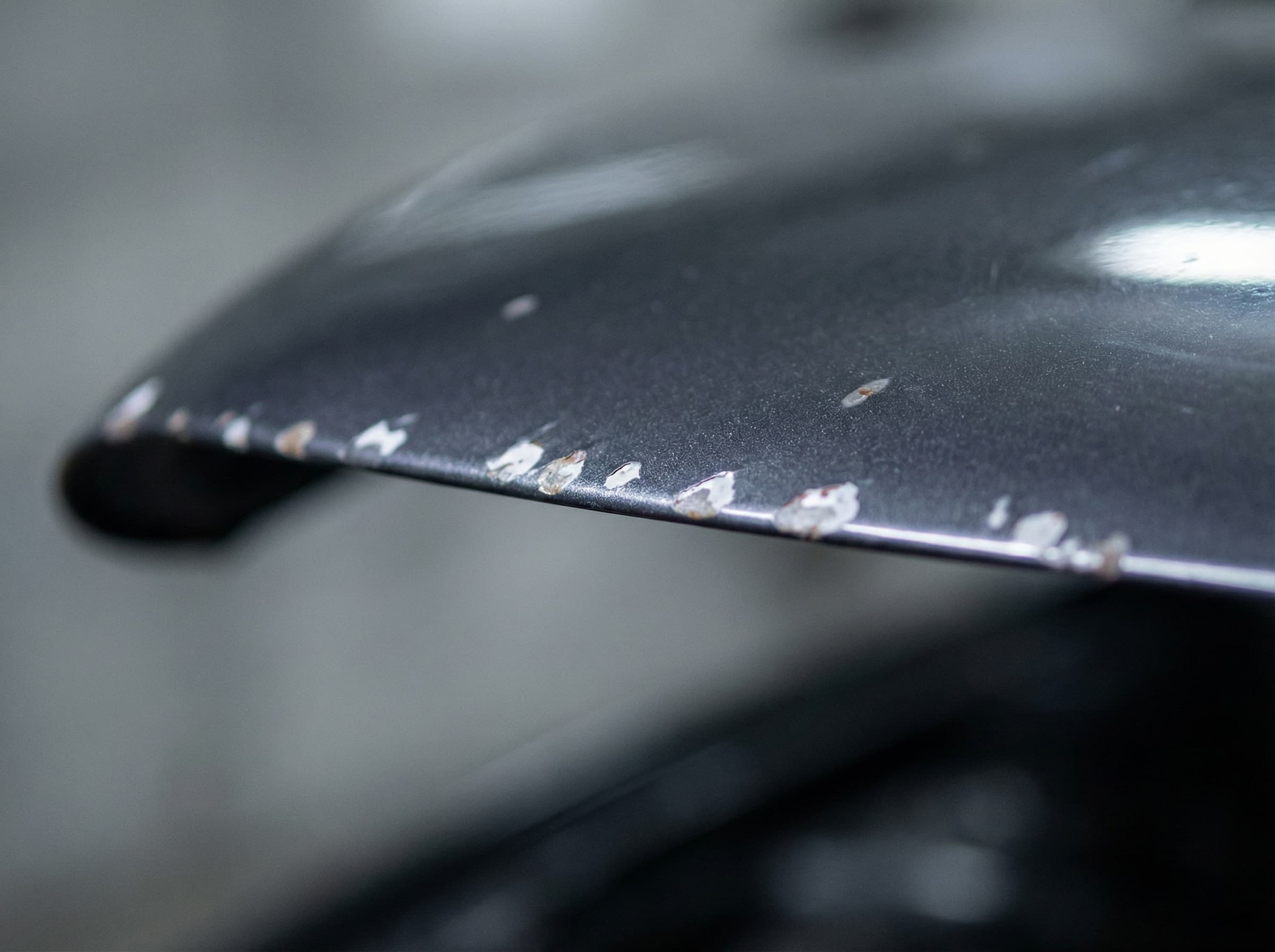 Close-up of stone chip damage on a dark car bonnet leading edge exposing primer underneath